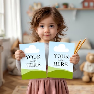 May include: Two rectangular paper sheets featuring a light blue sky, a white cloud, and a green grassy hill at the bottom. The text "YOUR design HERE" is printed in black on each sheet. Two yellow pencils are held alongside the right paper sheet.