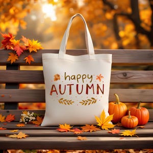May include: Cream-colored tote bag with the words "happy AUTUMN" in brown and orange. The bag is on a wooden bench with pumpkins and fall leaves. The background is a blurred view of autumn foliage.