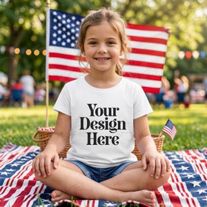 May include: A white short-sleeved t-shirt featuring the black text "Your Design Here" printed on the front. The shirt is worn by a child sitting on a blanket with a red, white, and blue star-spangled pattern, with a large American flag behind.