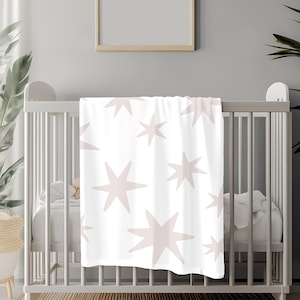 May include: A white crib with a pink and white star patterned blanket draped over the side. The crib is in a room with a white wall and a framed picture above it.
