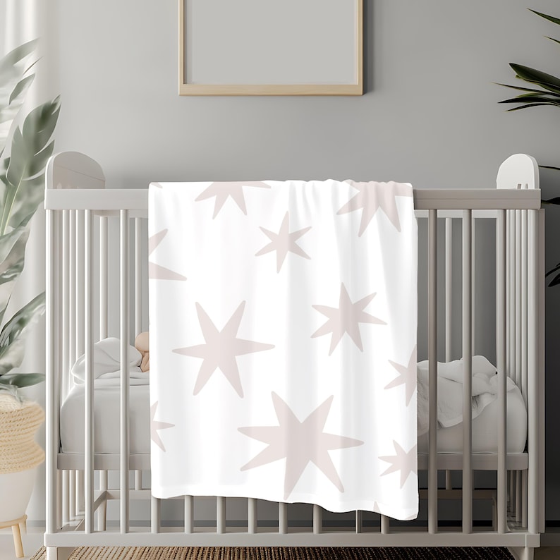 May include: A white crib with a pink and white star patterned blanket draped over the side. The crib is in a room with a white wall and a framed picture above it.