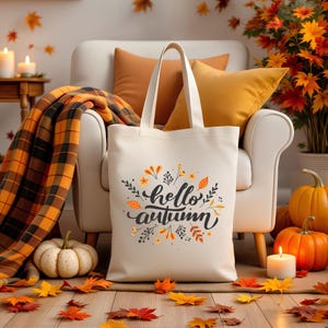 May include: A cream-colored tote bag with the words "hello autumn" in black script, surrounded by floral and leaf designs in orange and yellow. The bag is set against a cozy autumn scene with pumpkins, candles, and a plaid blanket.