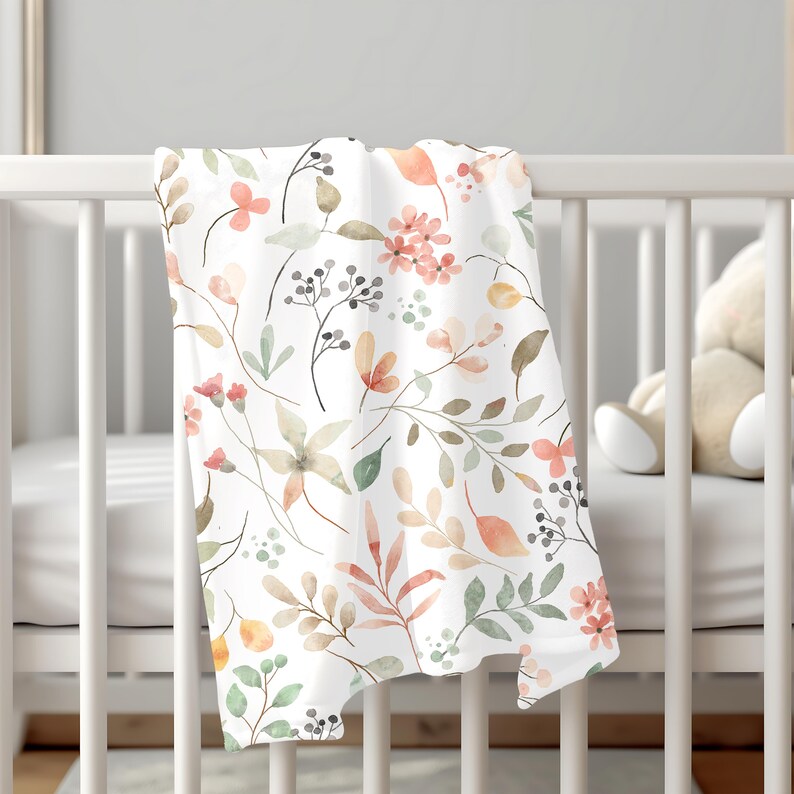 K&ouml;nnte beinhalten: Eine wei&szlig;e Babydecke mit einem Aquarell-Blumenmuster mit rosa, orangefarbenen und gr&uuml;nen Blumen und Bl&auml;ttern.