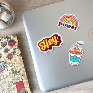 May include: A collection of colorful stickers on a laptop. The stickers include a rainbow with the word "power", the word "Hey" in yellow script, and a cartoon iced coffee. A decorative diary and a glass bowl with seashells are also visible.