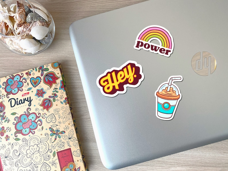 May include: A collection of colorful stickers on a laptop. The stickers include a rainbow with the word "power", the word "Hey" in yellow script, and a cartoon iced coffee. A decorative diary and a glass bowl with seashells are also visible.