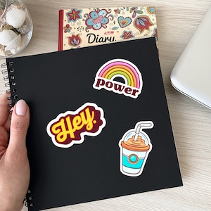 May include: A black spiral-bound notebook with three colorful stickers: a rainbow with the word "power", the word "Hey" in a yellow font, and a cartoon drink. A diary and seashells are in the background.