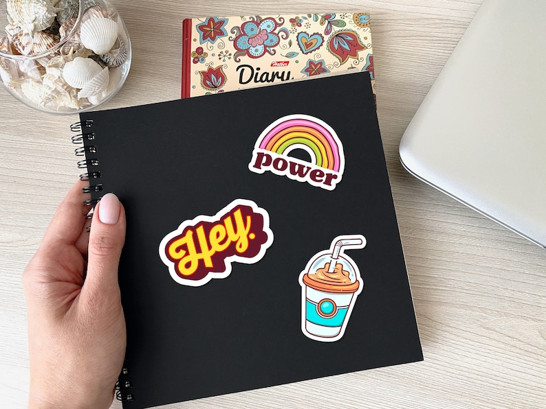 May include: A black spiral-bound notebook with three colorful stickers: a rainbow with the word "power", the word "Hey" in a yellow font, and a cartoon drink. A diary and seashells are in the background.