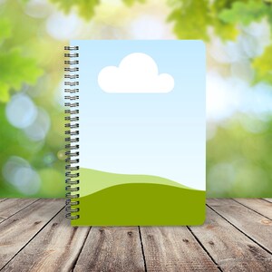 May include: A spiral bound notebook with a green and blue cover featuring a white cloud and a green hill. The notebook is on a wooden surface.