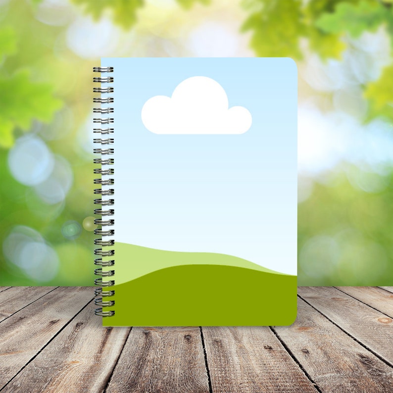 May include: A spiral bound notebook with a green and blue cover featuring a white cloud and a green hill. The notebook is on a wooden surface.