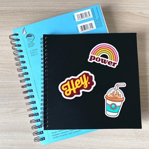 May include: Two spiral-bound sketchbooks, one black and one light blue, are stacked. The black sketchbook has stickers that say "Hey", "power", and a drink. The light blue sketchbook is partially visible.