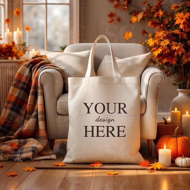 May include: A cream-colored tote bag with the text "YOUR design HERE" in black. The bag is in front of a cream-colored armchair, with a plaid blanket, pumpkins, candles, and fall foliage in the background.