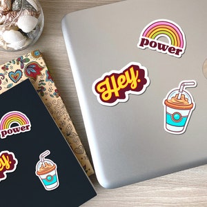 May include: A collection of colorful stickers featuring a rainbow with the word "power", the word "Hey!", and a cartoon iced coffee. The stickers are on a laptop and a notebook with floral designs.