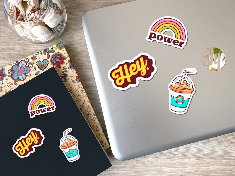 May include: A collection of colorful stickers featuring a rainbow with the word "power", the word "Hey!", and a cartoon iced coffee. The stickers are on a laptop and a notebook with floral designs.