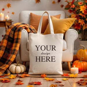 May include: A cream-colored tote bag with the text "YOUR design HERE" in black, standing in front of a white armchair with orange pillows. A plaid blanket, pumpkins, and fall leaves complete the autumn-themed scene.