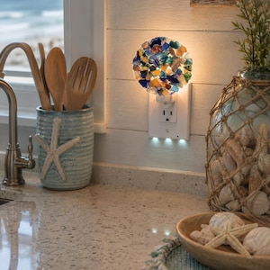 May include: A circular night light plugged into a wall outlet, featuring a mosaic of multi-colored glass pieces in shades of blue, green, amber, and clear. The light emits a warm glow, illuminating the textured glass fragments from behind.