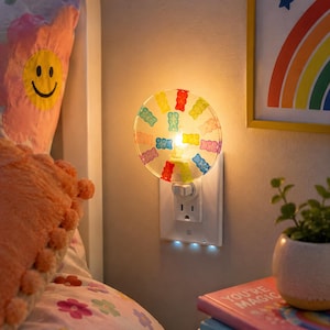 May include: A circular plug-in night light featuring a clear, transparent disc decorated with colorful, translucent gummy bear-shaped accents in red, yellow, green, blue, and purple, emitting a warm, soft glow from the center of the fixture.