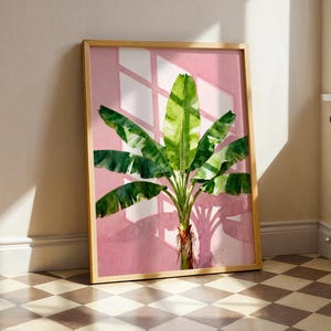 May include: A framed watercolour print of a banana tree with large green leaves and a pink background. The artwork is in a light wood frame and leans against a wall. The shadow of a window is cast on the pink background.