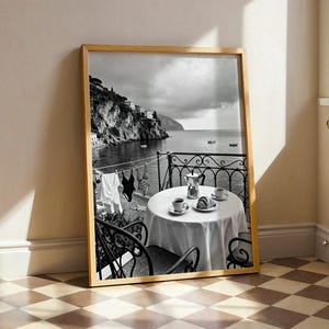 May include: A framed black and white print featuring a balcony overlooking a coastal landscape. A round table with a white tablecloth holds a coffee pot, two cups, and a croissant. Swimwear hangs on a clothesline against a sea view.