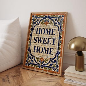 Puede incluir: Una obra de arte en mosaico enmarcada con las palabras "HOME SWEET HOME" en letras azules. El mosaico presenta un diseño floral con detalles azules, naranjas y verdes. El marco es de madera clara.