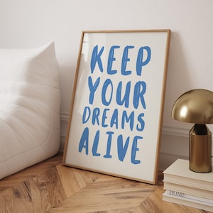May include: A framed print with the text "KEEP YOUR DREAMS ALIVE" in blue lettering on a white background. The print is sitting on a wooden floor with a gold lamp and books.