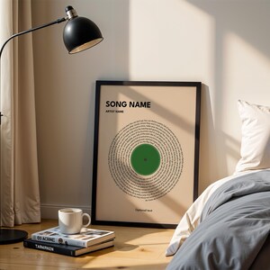 May include: A framed print with a circular design resembling a vinyl record. The print features a green center and text arranged in concentric circles. The words "SONG NAME" and "ARTIST NAME" are at the top. A black floor lamp and a white mug are also visible.