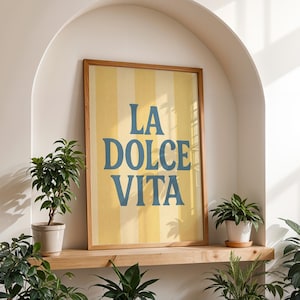 May include: A framed art print with the words "LA DOLCE VITA" in blue lettering on a yellow and white striped background. The print is displayed in a wooden frame. The artwork is set in a white alcove with potted plants on a wooden shelf.