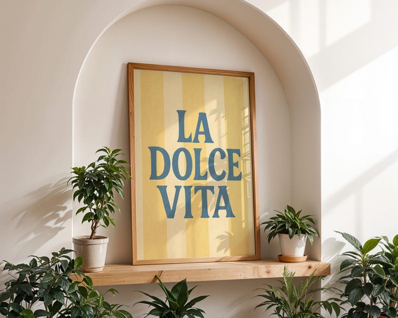 May include: A framed art print with the words "LA DOLCE VITA" in blue lettering on a yellow and white striped background. The print is displayed in a wooden frame. The artwork is set in a white alcove with potted plants on a wooden shelf.