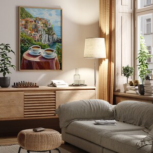 May include: A painting of a colorful Italian village on a cliff overlooking the ocean. Two cups of coffee are on a table in the foreground.