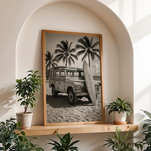 May include: Black and white framed print of a vintage Land Rover parked on a sandy beach with a surfboard leaning against it. Palm trees are in the background, evoking a tropical vacation theme.
