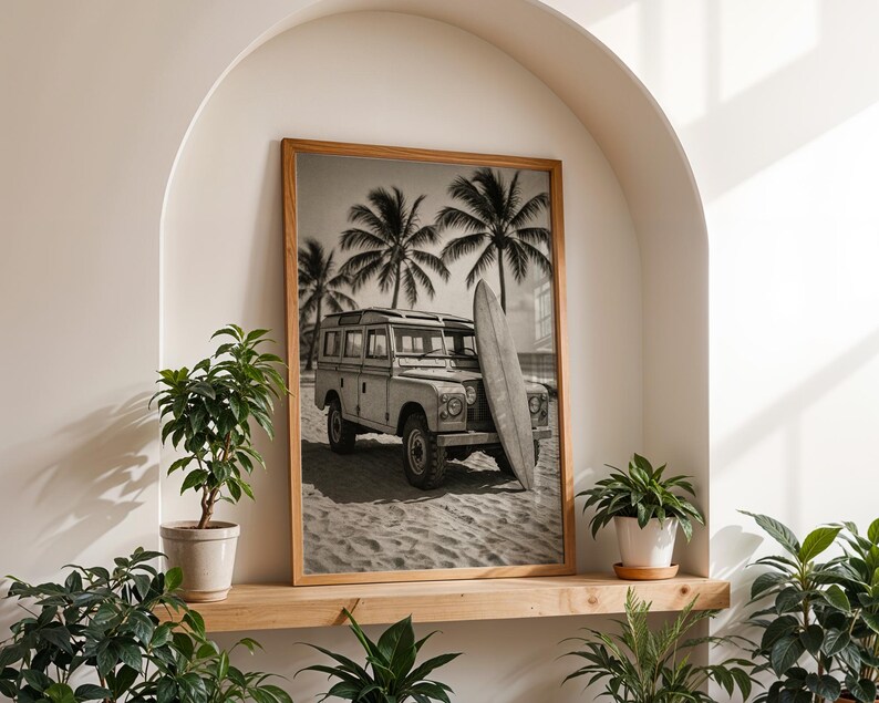 May include: Black and white framed print of a vintage Land Rover parked on a sandy beach with a surfboard leaning against it. Palm trees are in the background, evoking a tropical vacation theme.