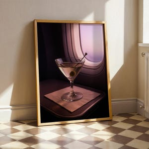 May include: A framed print featuring a martini in a stemmed glass with an olive on a toothpick. The drink sits on a small napkin on a tray, with an airplane window in the background. The frame is a light wood color.
