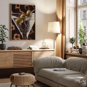 May include: A living room interior with a light brown couch, a wooden side table, a floor lamp with a white lampshade, and a framed print of a martini glass with olives. The print has a brown background with a leopard print pattern.