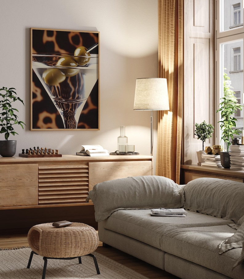 May include: A living room interior with a light brown couch, a wooden side table, a floor lamp with a white lampshade, and a framed print of a martini glass with olives. The print has a brown background with a leopard print pattern.