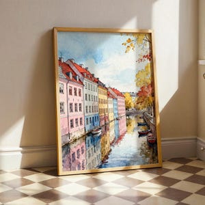 May include: Watercolor painting of a European canal scene. Colorful buildings line the water's edge, reflecting in the calm water. The buildings are painted in shades of pink, yellow, and blue, with red rooftops. Small boats are docked along the canal.