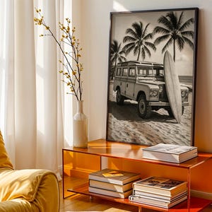 May include: Black and white framed print of a vintage SUV parked on a sandy beach with a surfboard. Palm trees are in the background. The print is displayed on an orange console table with books and a vase with yellow flowers.