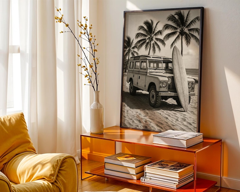 May include: Black and white framed print of a vintage SUV parked on a sandy beach with a surfboard. Palm trees are in the background. The print is displayed on an orange console table with books and a vase with yellow flowers.