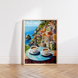 May include: A painting of a colorful Italian village on a cliff overlooking the ocean. Two cups of coffee are on a blue table in the foreground.