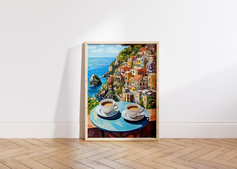May include: A painting of a colorful Italian village on a cliff overlooking the ocean. Two cups of coffee are on a blue table in the foreground.