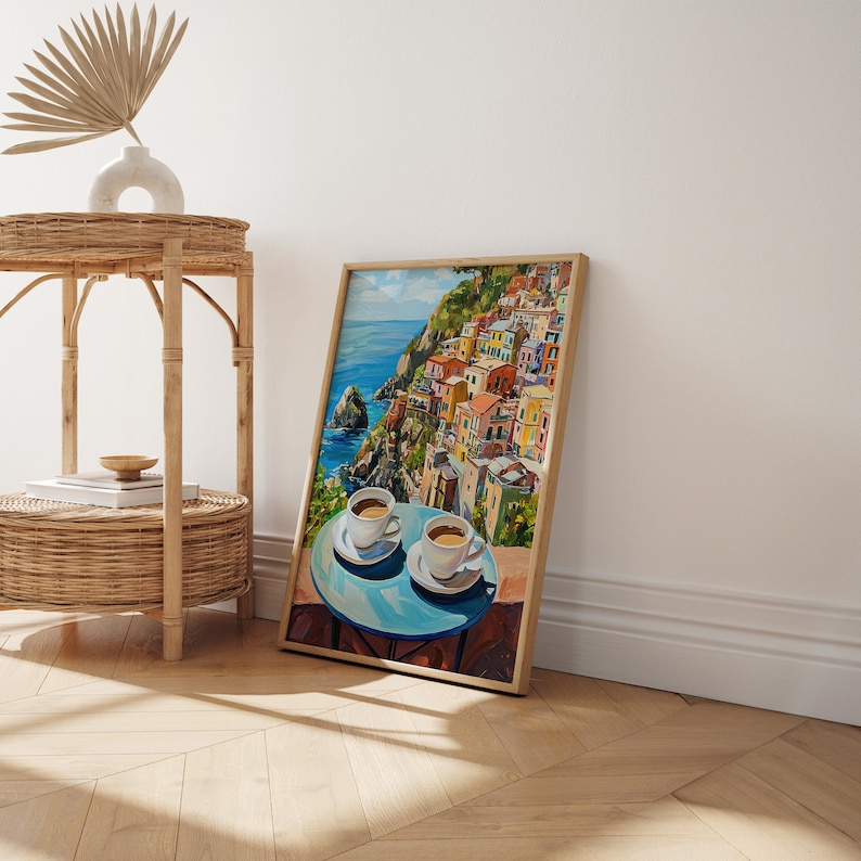May include: A framed painting of a colorful Italian village on a cliff overlooking the ocean. Two cups of coffee are on a blue table in the foreground.