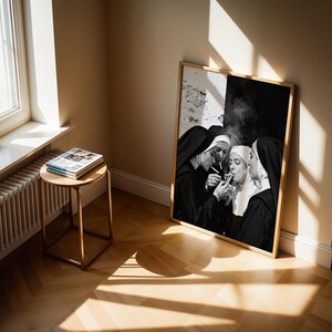 May include: A framed black and white photograph depicts four nuns in traditional habits, gathered closely together, lighting cigarettes. The artwork is leaning against a wall in a room with a window and a small side table with books.