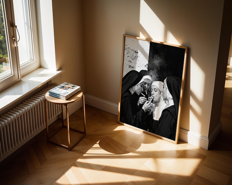 May include: A framed black and white photograph depicts four nuns in traditional habits, gathered closely together, lighting cigarettes. The artwork is leaning against a wall in a room with a window and a small side table with books.