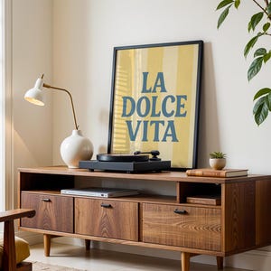 May include: A mid-century modern style wooden media console with three drawers and a shelf. A framed poster with the text "LA DOLCE VITA" is displayed on top, along with a record player and a white lamp.
