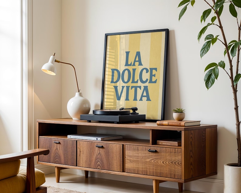 May include: A mid-century modern style wooden media console with three drawers and a shelf. A framed poster with the text "LA DOLCE VITA" is displayed on top, along with a record player and a white lamp.
