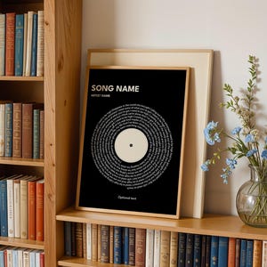 May include: A framed black print with white text arranged in a circular pattern, resembling a vinyl record. The print includes the text "SONG NAME" and "ARTIST NAME" at the top, with "Optional text" at the bottom. The print is displayed on a wooden shelf with books.