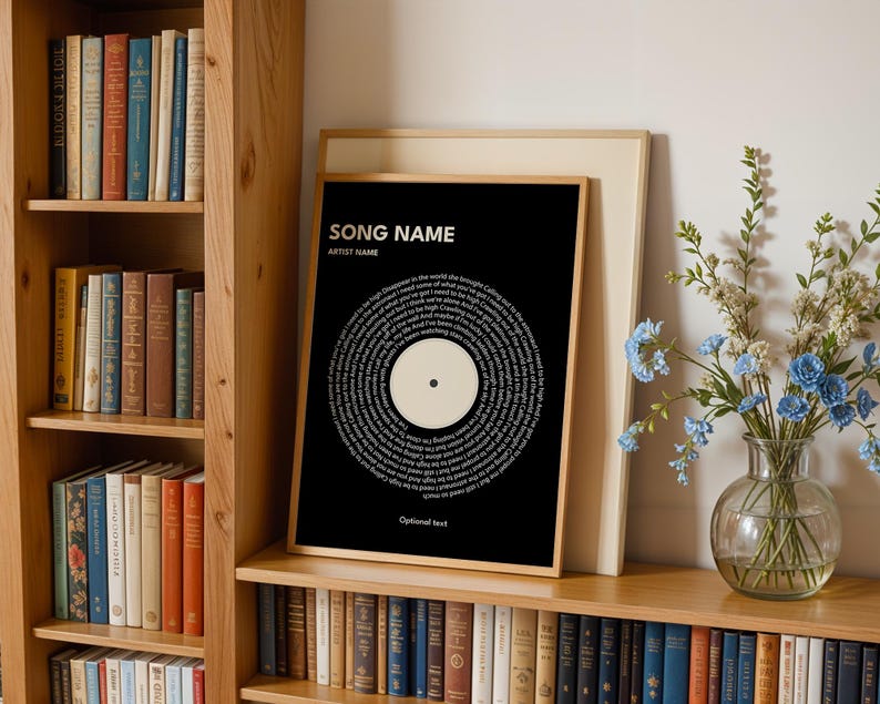May include: A framed black print with white text arranged in a circular pattern, resembling a vinyl record. The print includes the text "SONG NAME" and "ARTIST NAME" at the top, with "Optional text" at the bottom. The print is displayed on a wooden shelf with books.