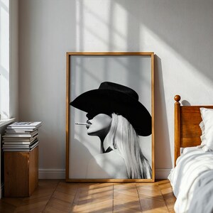 May include: Black and white framed art print featuring a woman in a wide-brimmed hat, smoking a cigarette. The artwork has a minimalist style with a focus on the woman's profile and long, flowing hair. The print is displayed in a room with natural light.
