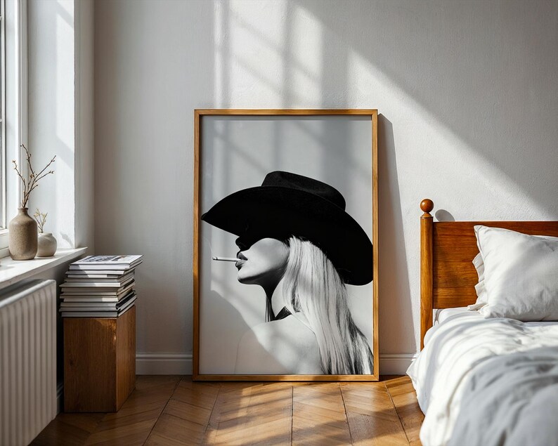 May include: Black and white framed art print featuring a woman in a wide-brimmed hat, smoking a cigarette. The artwork has a minimalist style with a focus on the woman's profile and long, flowing hair. The print is displayed in a room with natural light.