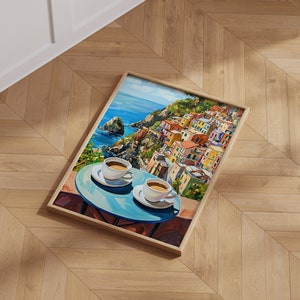May include: A framed painting of a colorful Italian village on a cliffside overlooking the ocean. Two cups of coffee are on a blue table in the foreground.