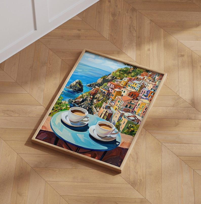 May include: A framed painting of a colorful Italian village on a cliffside overlooking the ocean. Two cups of coffee are on a blue table in the foreground.