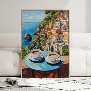 May include: A painting of a colorful Italian village on a cliff overlooking the ocean. Two cups of coffee sit on a blue table in the foreground.